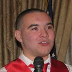 Man speaking into microphone wearing red sash